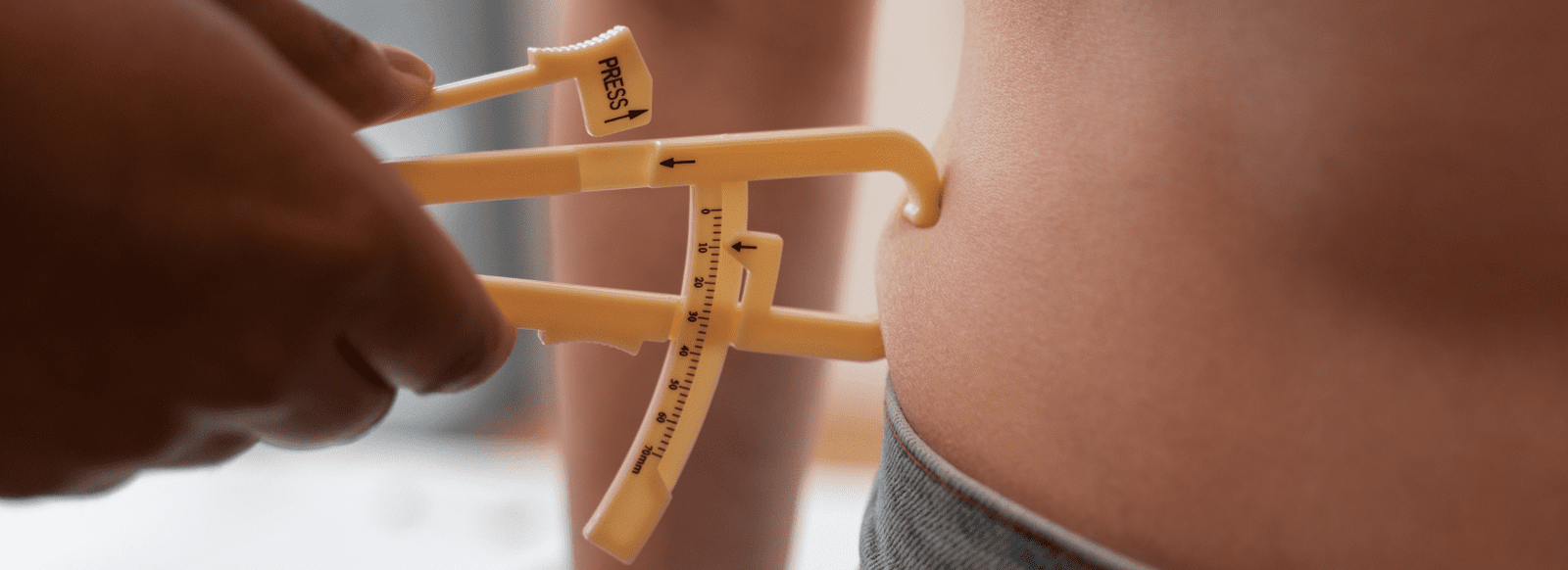 Close-up of a person using calipers to measure body fat on the side of someone's abdomen, with the person wearing jeans and a cropped top.